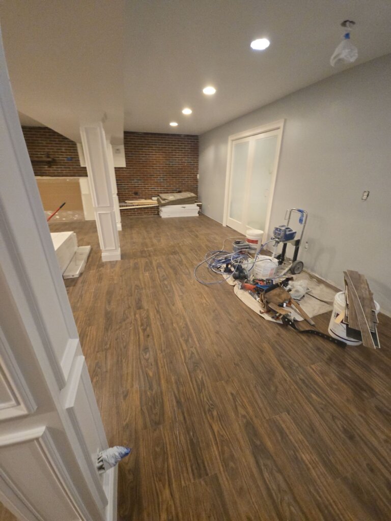 Interior custom home build progress by Redstone Ridge Homes, showing new wood-look flooring, grey walls, a brick accent wall, and construction tools.