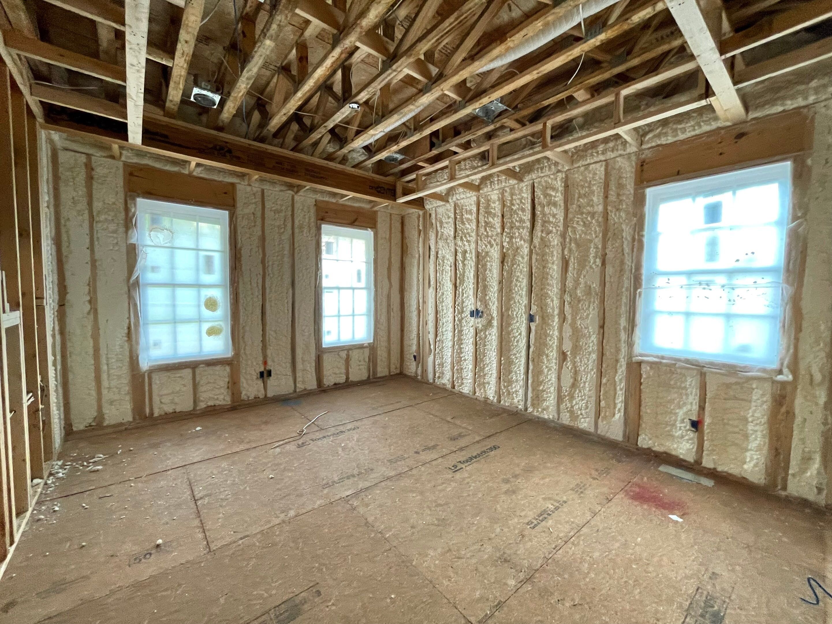 Residential spray foam insulation applied between wall studs in a new home construction project with open ceiling framing and OSB subflooring.