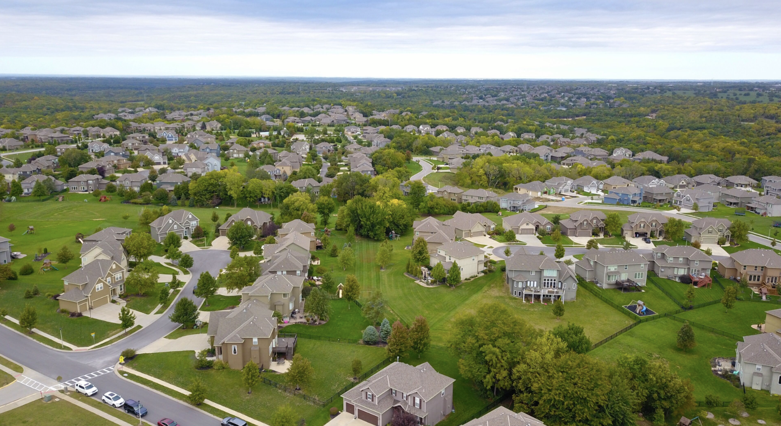 An aerial, high-angle view of a sprawling suburban neighborhood in North Alabama, featuring multi-story single-family homes with large green lawns, winding streets, and a dense treeline stretching into the horizon under an overcast sky.


