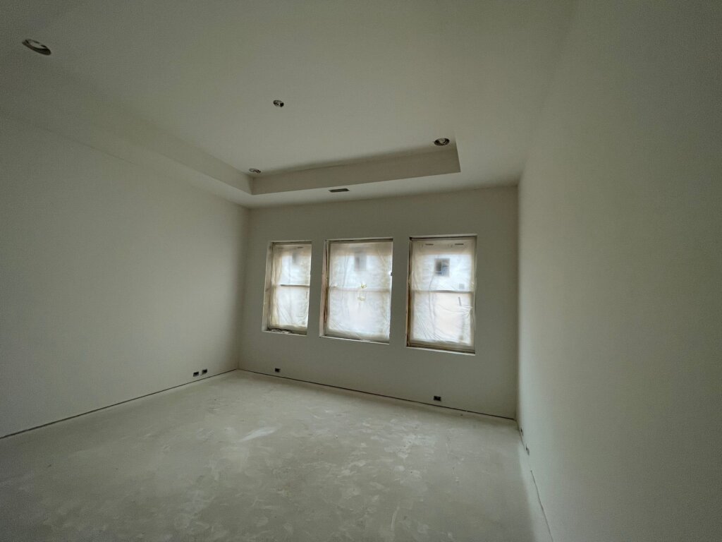 Freshly painted off-white bedroom featuring a coffered tray ceiling, recessed lighting cutouts, and three plastic-covered windows during renovation.