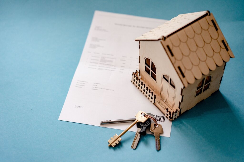 Image of a small wooden house upon a stack of paperwork, beside a ring of housekeys.